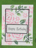 A handmade square greeting card with a pink and green floral pattern, featuring a central black-bordered label that reads 'Happy Birthday' in black script. The card is photographed flat against a green glittery background.
