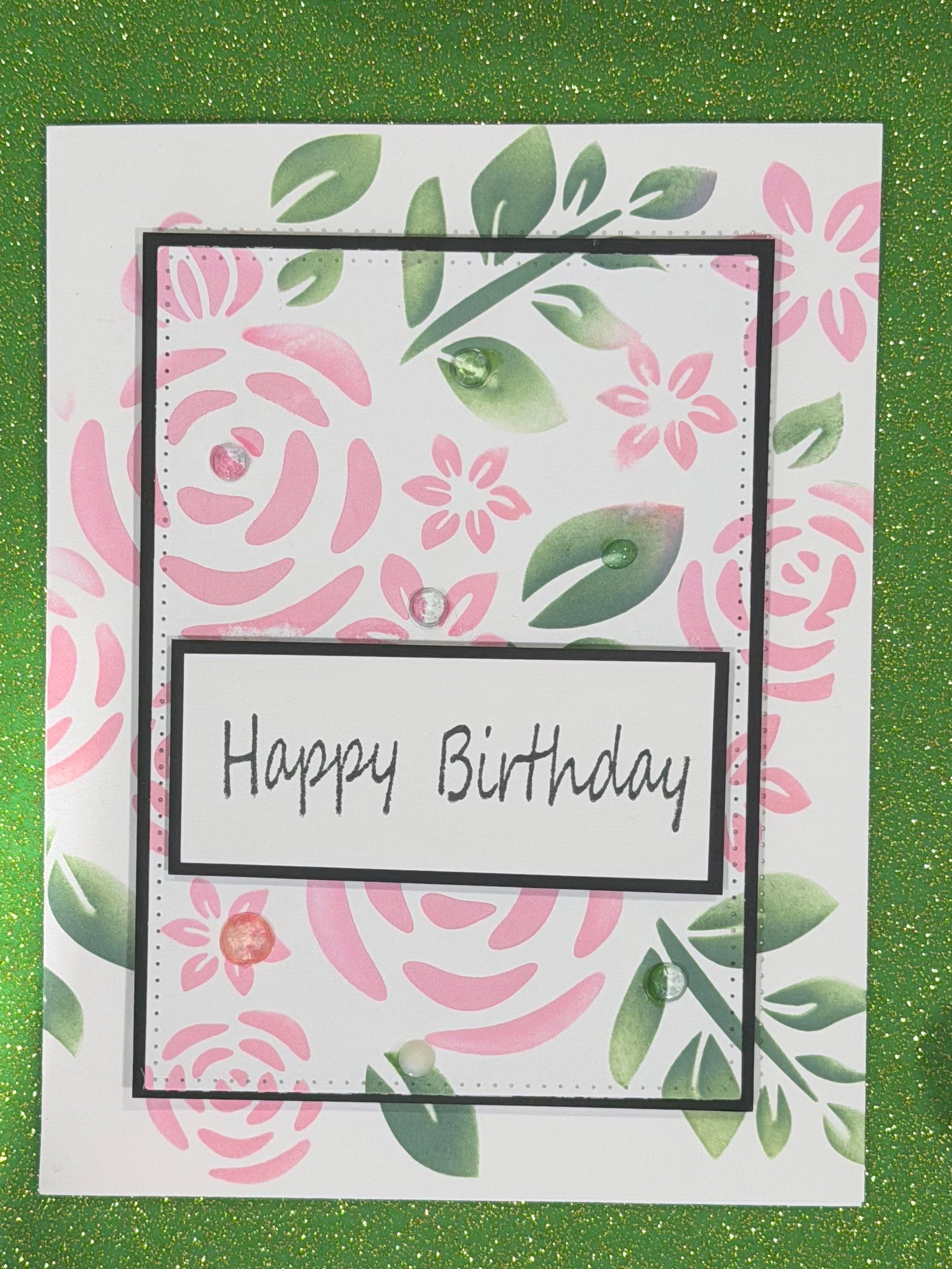 A handmade square greeting card with a pink and green floral pattern, featuring a central black-bordered label that reads 'Happy Birthday' in black script. The card is photographed flat against a green glittery background.