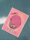 A pink 'Get Well Soon' greeting card with a glittery circular center and a small pink bow, placed on a dark blue glittery surface.