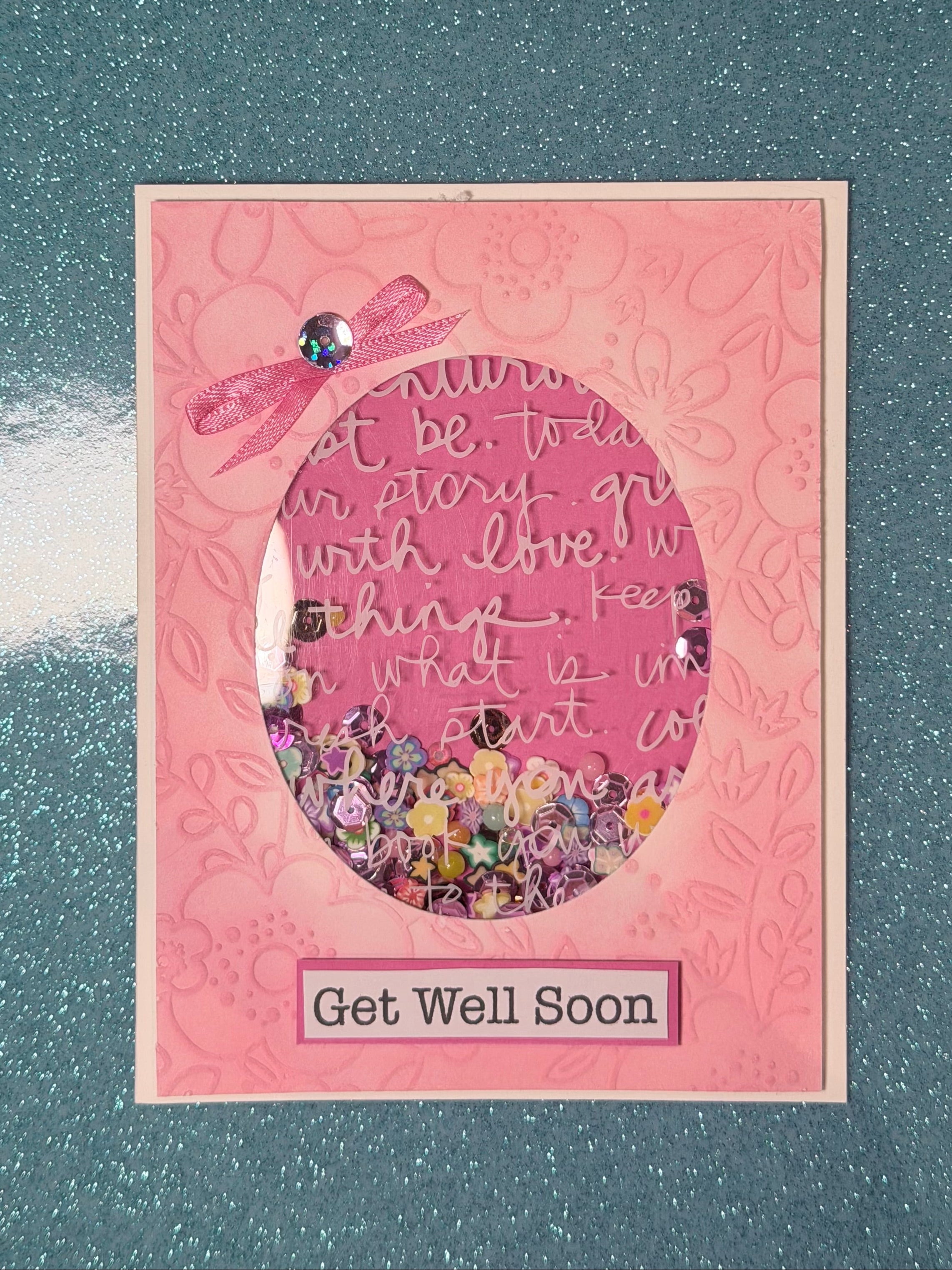 A handmade pink 'Get Well Soon' card featuring a glittery pink circular shaker filled with confetti and rhinestones, placed on a dark teal glittery surface.