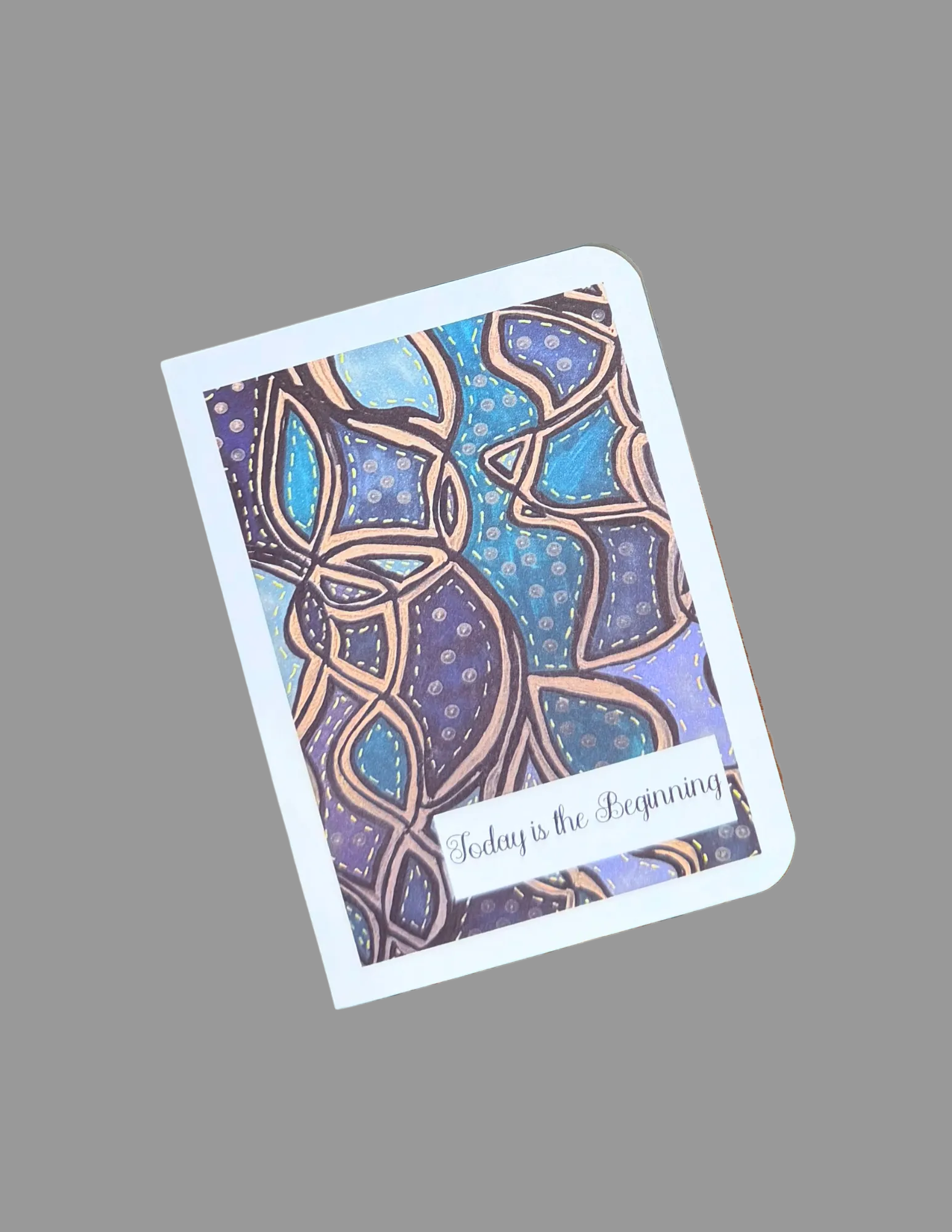 A rectangular art print with rounded corners lies on a brown wooden surface. The print features an abstract, geometric pattern in shades of blue, purple, and gold, with a small white banner at the bottom containing the text 'Today is the Beginning'.